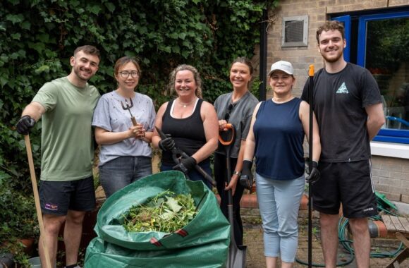 Volunteers from JTI standing in a line holding gardening tools and a bag of garden waste after a volunteering session in the outdoor area of a Look Ahead mental health service.