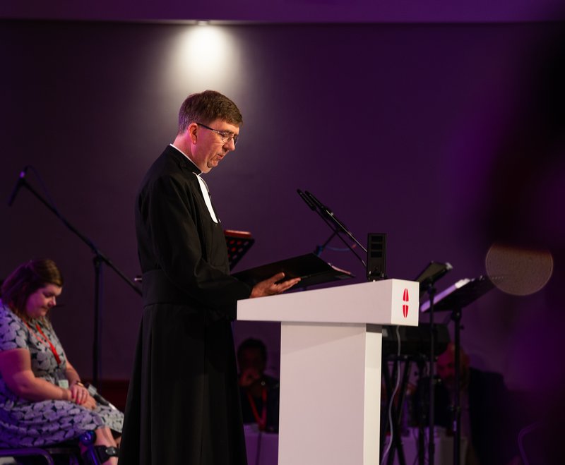 A side-on shot of Jonathan Hustler speaking at the Methodist Conference from a podium at the front of the hall.